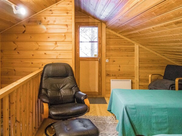 A wooden cabin interior with a single leather chair, a table with a green tablecloth, and a window with a view of bare trees.