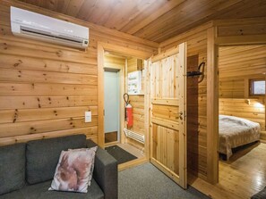 Ein Interieur einer Holzhütte mit einer Couch, einem Bett, einer Klimaanlage und einer Tür zu einem weiteren Raum.