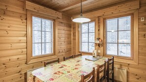Interior de madera con una mesa de comedor cubierta por un mantel colorido, flanqueada por ventanas con persianas, y un lámpara colgante.