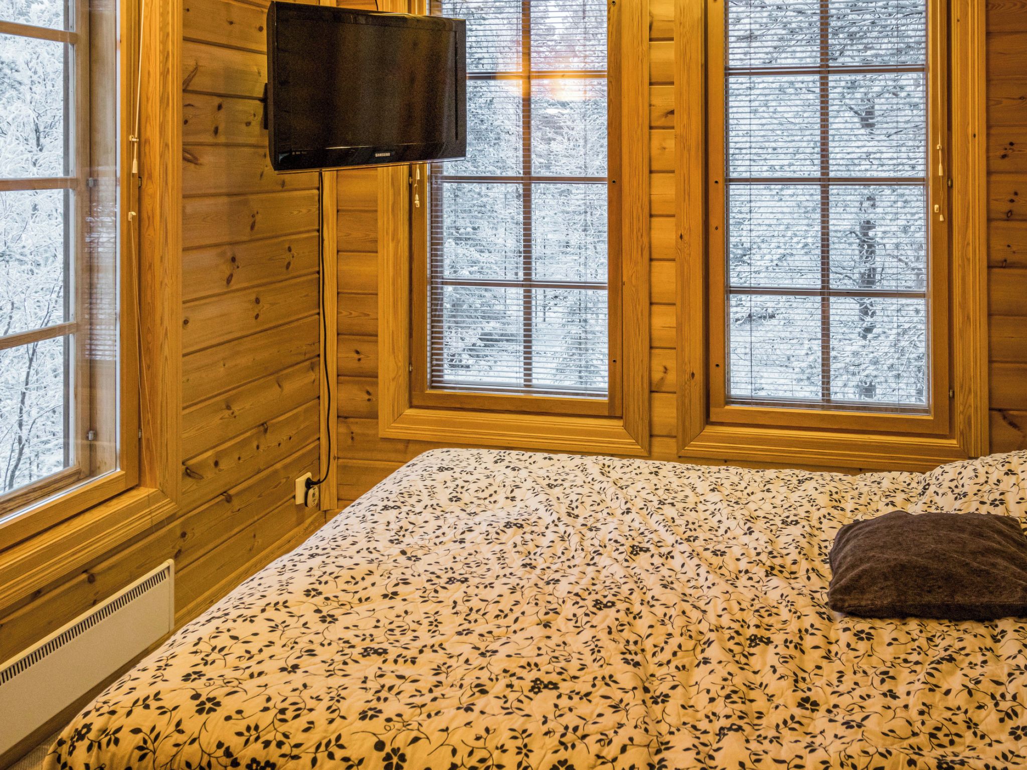 Une pièce aux murs en bois, un lit recouvert d’une couvre-lit à motifs floraux, un téléviseur fixé au mur et des fenêtres à vitres dépolies.