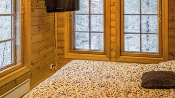 Une pièce aux murs en bois, un lit recouvert d’une couvre-lit à motifs floraux, un téléviseur fixé au mur et des fenêtres à vitres dépolies.