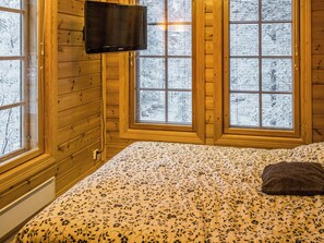A room with wooden walls, a bed with a floral bedspread, a television on the wall, and windows with frosted glass.