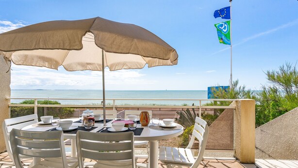 Ein Tisch für zwei Personen mit einem beigen Sonnenschirm, weißen Stühlen und Blick aufs Meer.