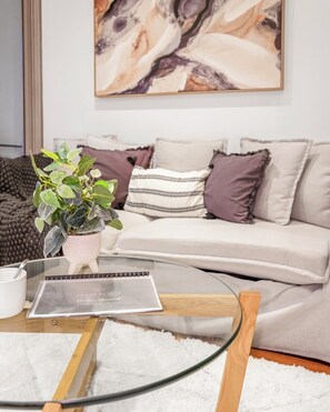 Ein modernes Wohnzimmer mit einem Glastisch, einer Topfpflanze, einem Notizbuch und einem Sofa mit gemusterten Kissen.