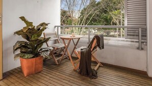 Uma varanda com planta em vaso, mesa com cadeira e vista para árvores e edifícios.