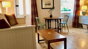 A cozy living room with a sofa, a wooden table, and two chairs. There is a window with curtains, a door, and a framed picture on the wall. - Beachfront Apartment With Woodburner & Courtyard (St Leonards-On-Sea)