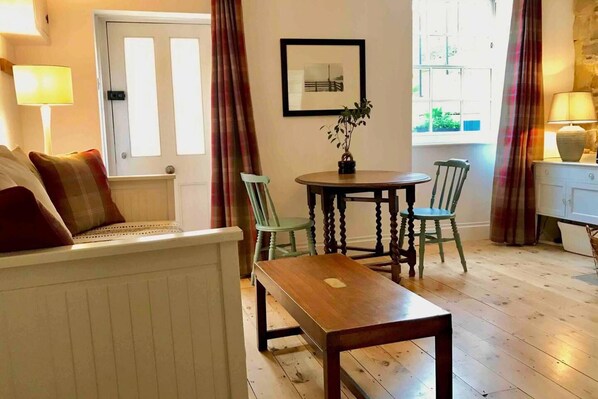 Un salón acogedor con un sofá, una mesa de madera y dos sillas. Se ve una ventana con cortinas, una puerta y un cuadro en la pared.