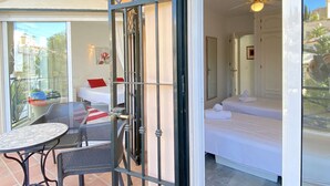 A room with a bed, a small table, and chairs, a balcony with a table and chairs, and a view of buildings outside.