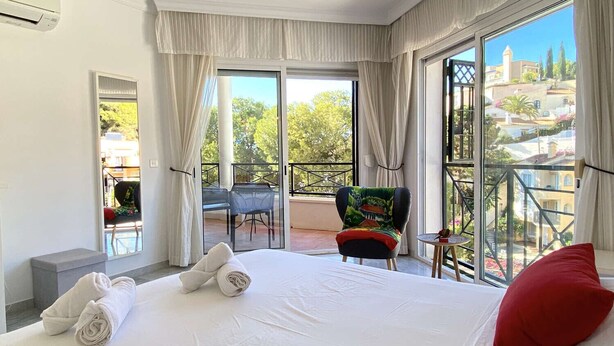 A hotel room with a large bed, a balcony with a chair, and a view of the city.