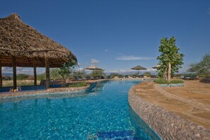 A swimming pool with clear blue water, surrounded by stone walls and shaded by thatched umbrellas.