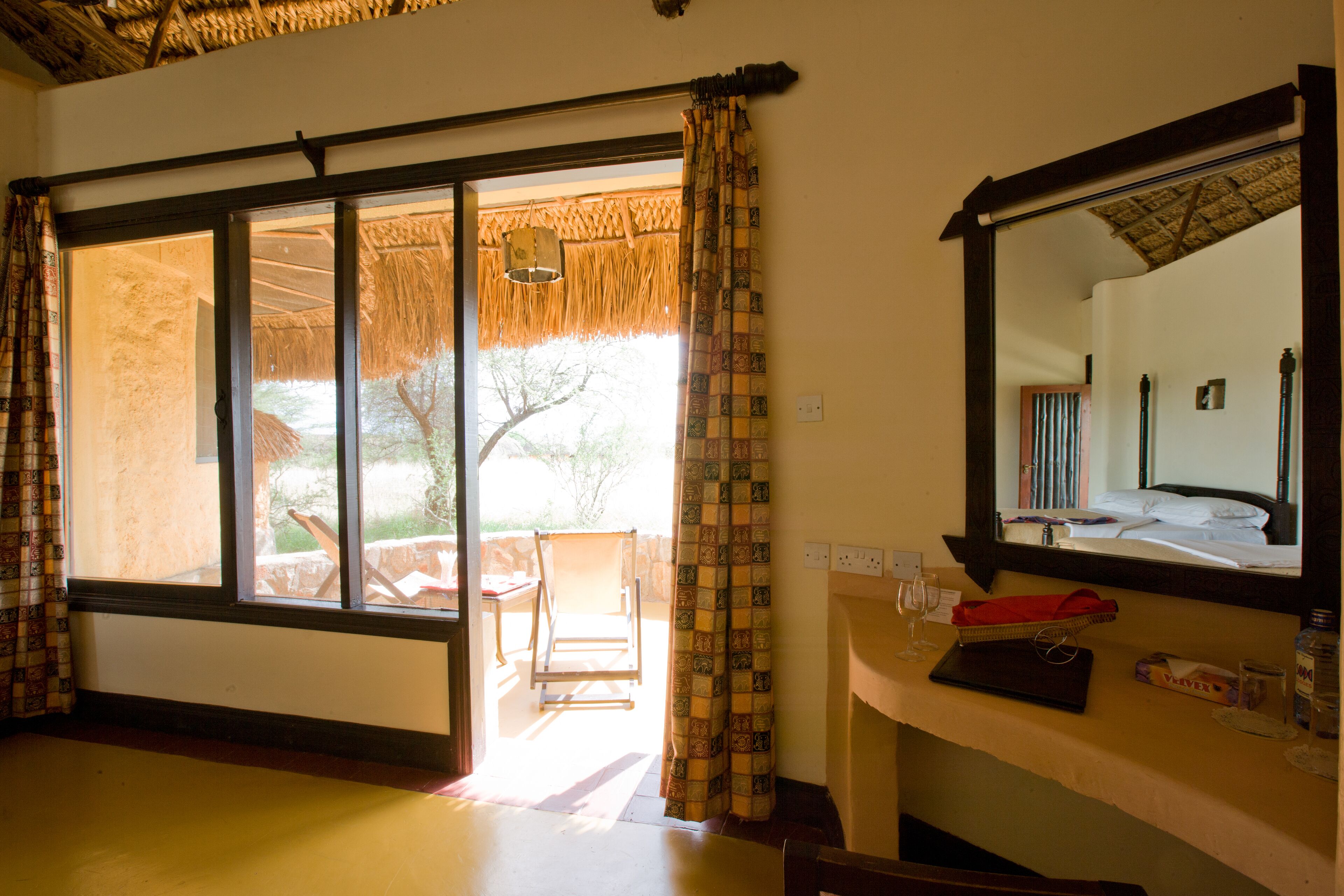 Habitación con puerta de vidrio corredera que da a un área exterior con una mesa y sillas, un espejo en la pared y un pequeño estante con una botella y un paño rojo.