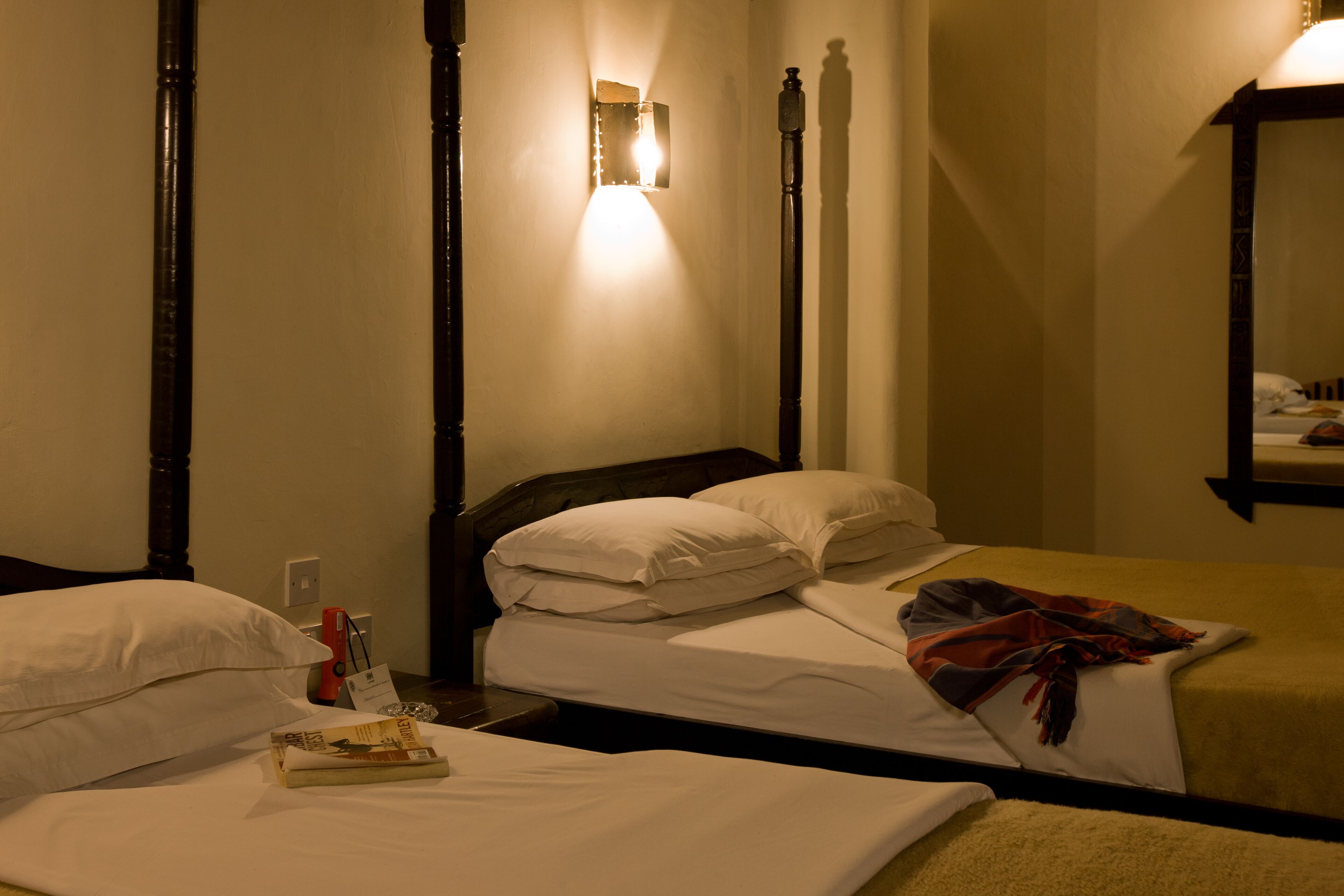 A hotel room with two beds, a nightstand, a bedside table, a wall-mounted lamp, and a mirror.