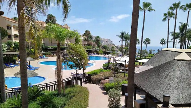Ein Resort mit mehreren Schwimmbecken, Palmen und einem Weg zu einem Gebäude mit einem Reetdach.