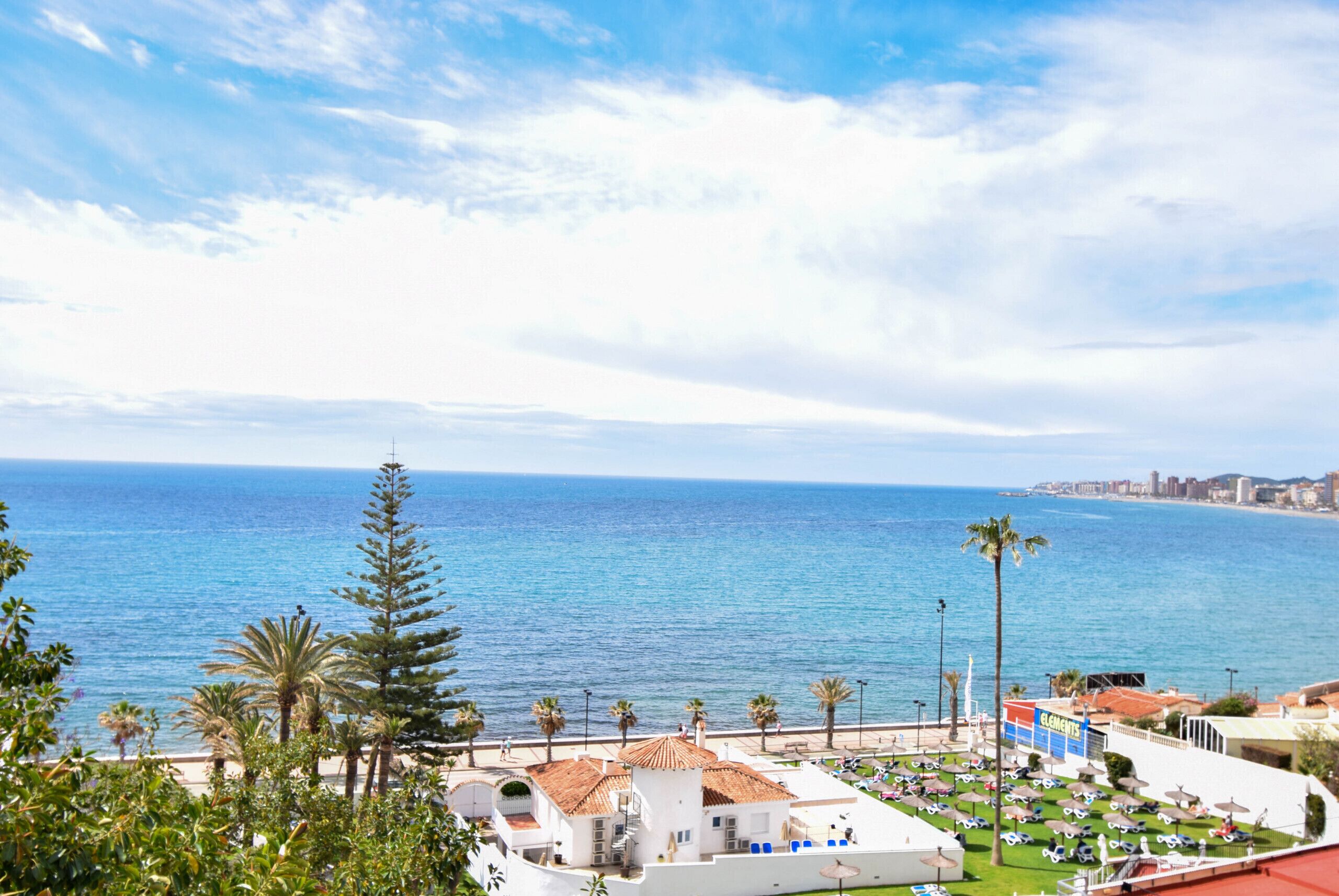 A coastal hotel with a view of the sea, palm trees, and a beach area with umbrellas.