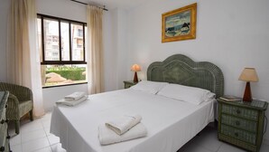 Ein Hotelzimmer mit einem Bett, zwei Nachttischen, einem Fenster mit Vorhängen, einem Korbsessel und einem Bild an der Wand.