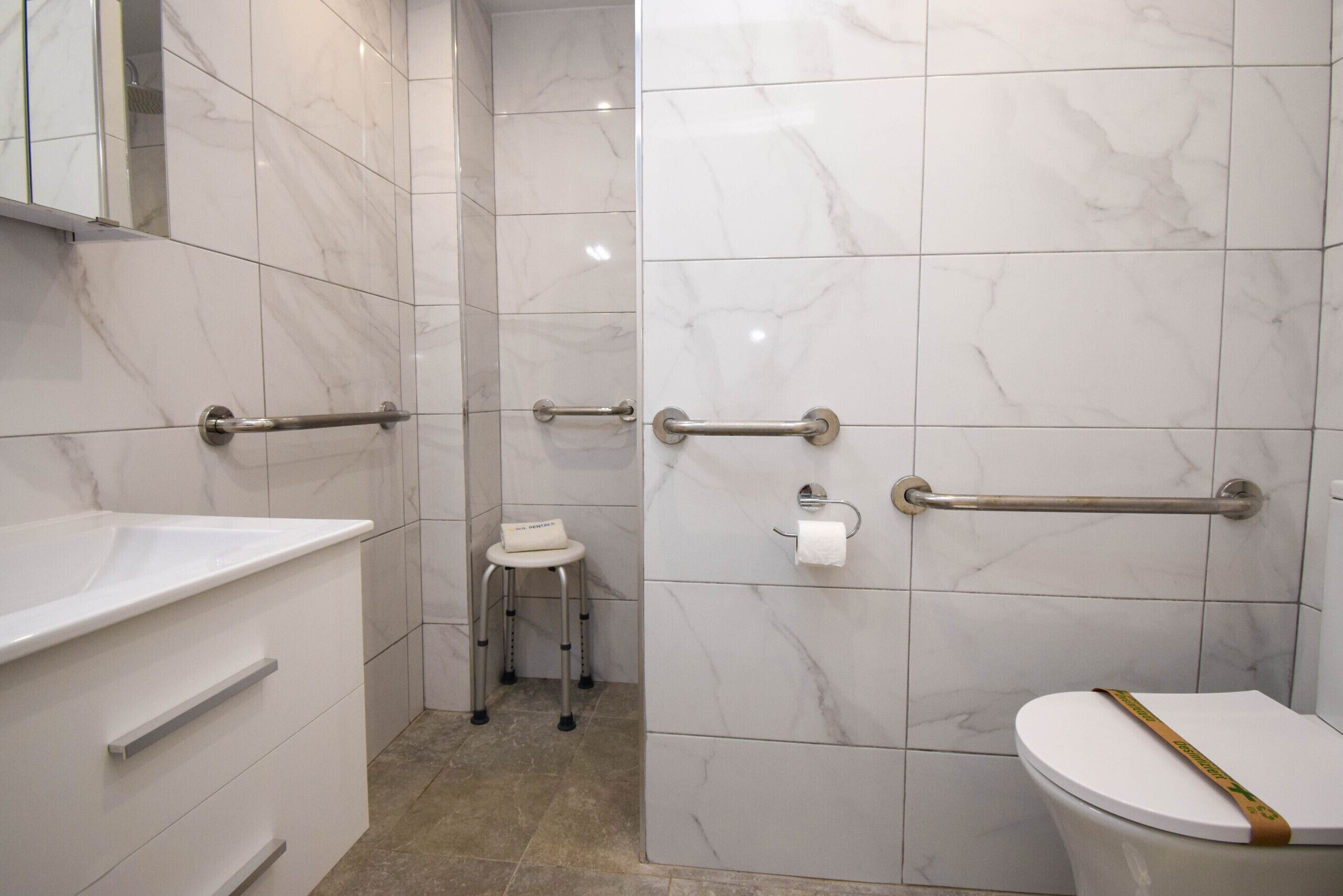 A bathroom with white marble tiles, a white toilet, a white sink, and a small stool.