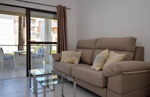 A living room with a beige sofa, a glass coffee table, and a view of a balcony with white chairs.