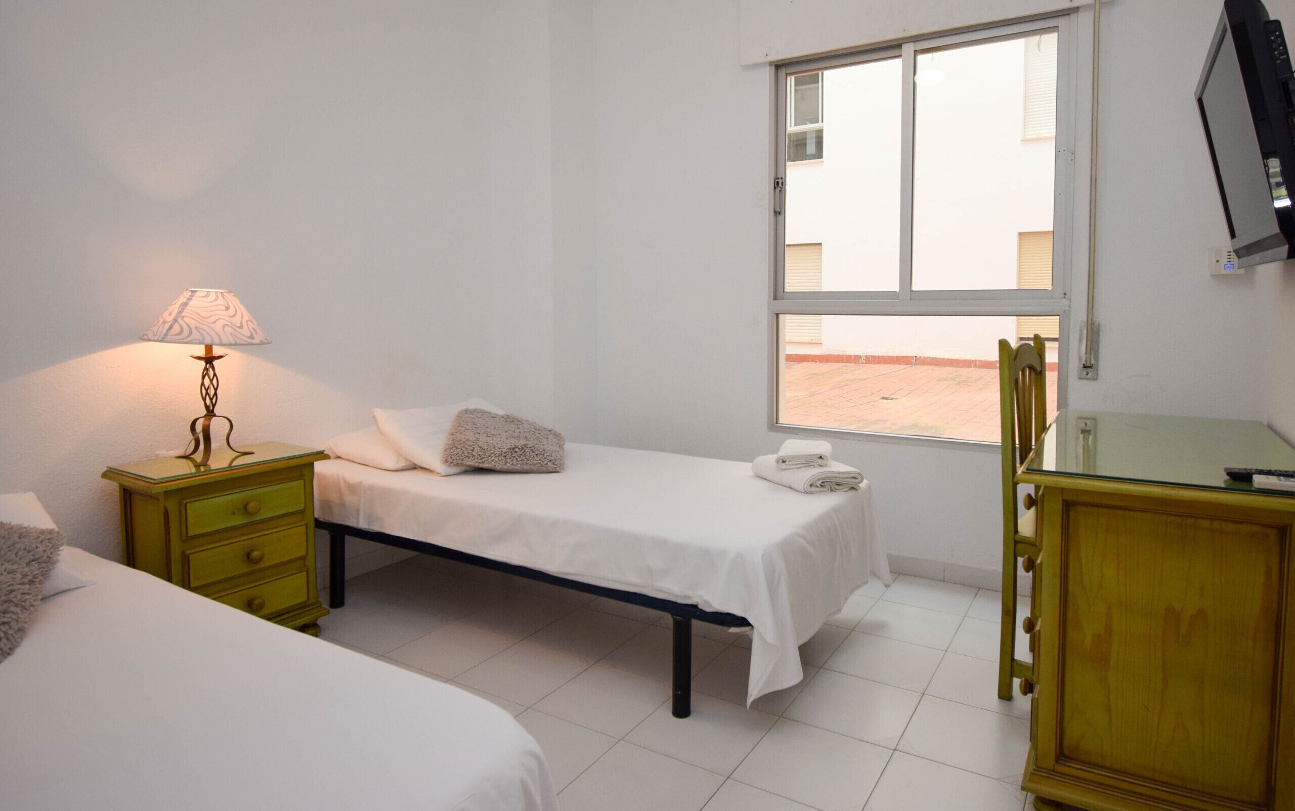 A bedroom with two beds, a bedside table with a lamp, a window, and a TV mounted on the wall.