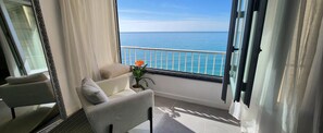 Ein Balkon mit Meerblick, ein Stuhl, ein kleiner Tisch mit Pflanze und ein Fenster mit Vorhängen.