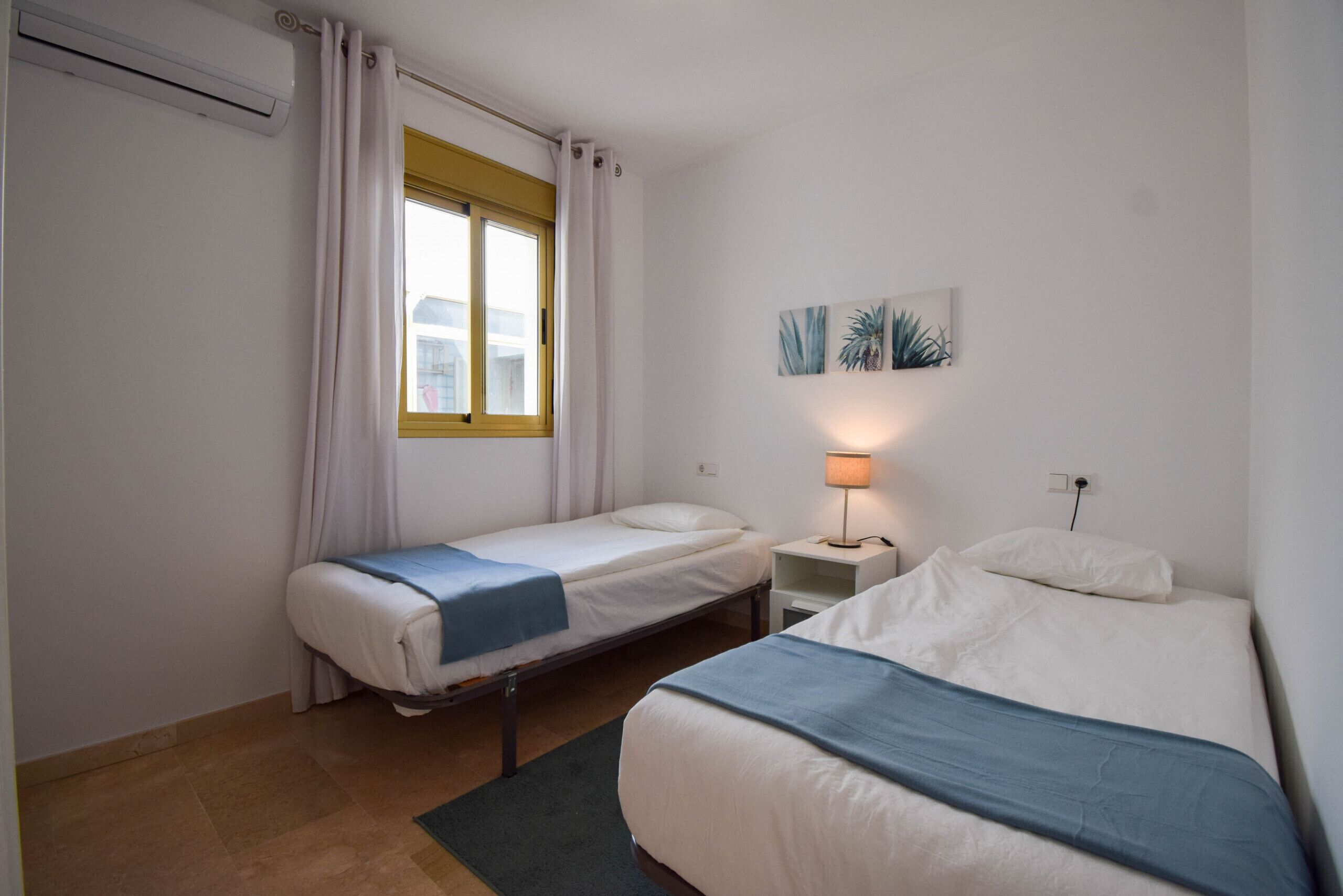 A small, clean room with two single beds, a window with curtains, a nightstand with a lamp, and a wall-mounted painting.