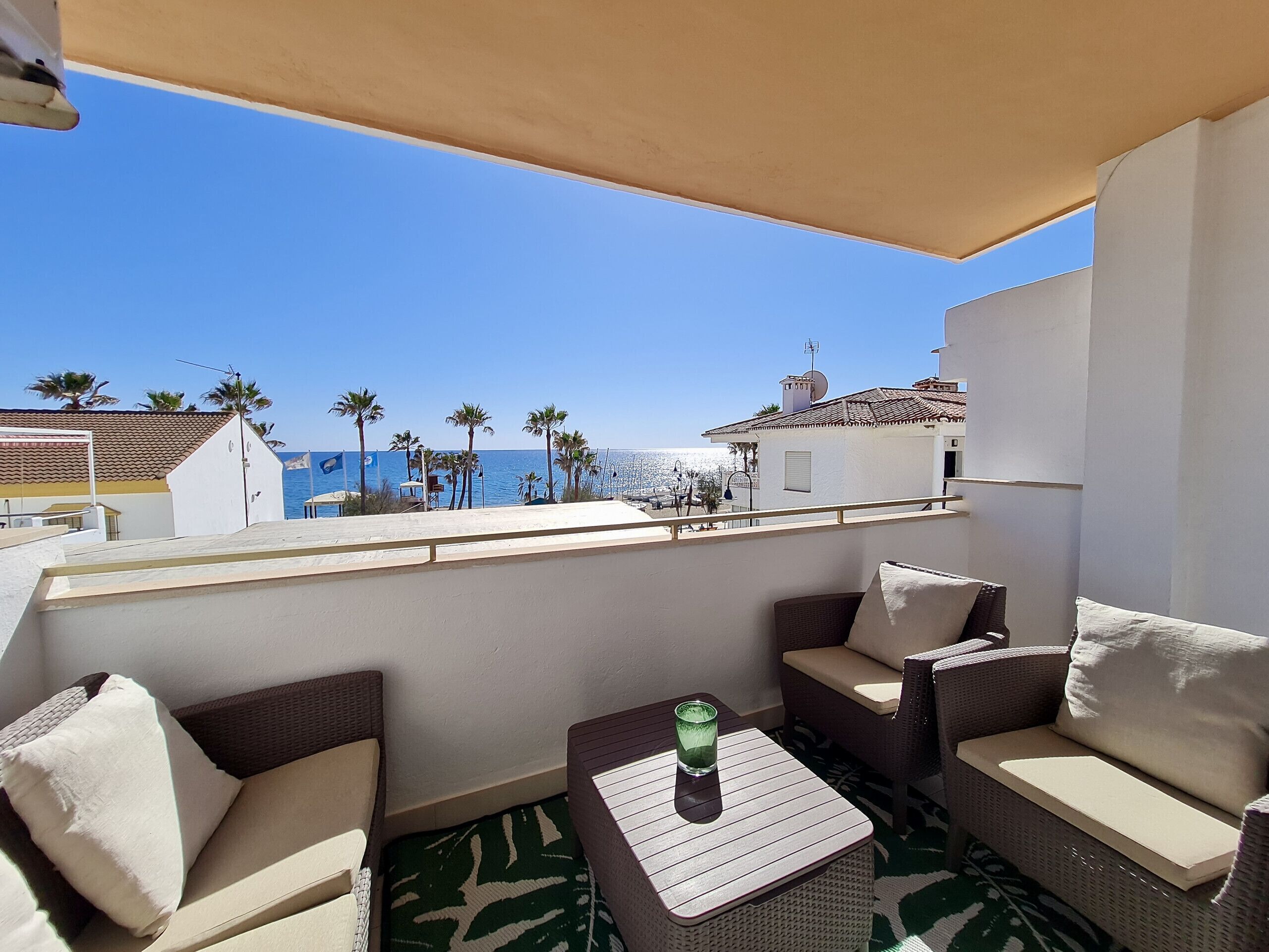A balcony with wicker furniture, a glass table, and a view of the sea and buildings.