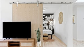 A modern living room with a large flat-screen TV on a wooden stand, a sliding wooden partition, a potted plant, a woven basket, and a black and white abstract wall art.
