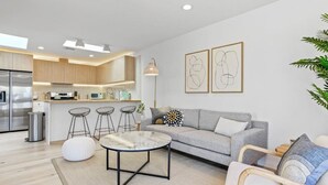 A modern kitchen with wooden cabinets, a built-in oven, and a stainless steel refrigerator. There is a dining area with a grey sofa, a round coffee table, and a bar stool. The room has a large window and a plant on the right.