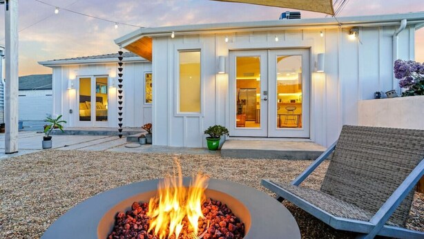 A modern house with a fire pit in the foreground, a covered patio area, and a view of the interior through the glass doors.