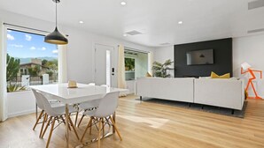 A modern living room with a white sofa, a dining table with chairs, and a TV mounted on the wall.