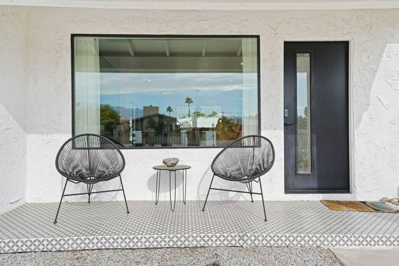 Un balcone con due sedie nere dal design a filo metallico, un tavolino rotondo con un oggetto decorativo e un'ampia finestra che si affaccia su edifici e palme.