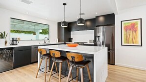 A modern kitchen with a central island, bar stools, stainless steel appliances, and a large window overlooking a green space.