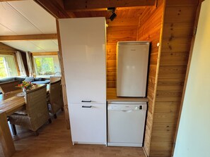 A compact kitchen with a refrigerator, a dishwasher, and a dining area with wicker chairs and a table. - Beachpark 2,Bungalow 62 (Renesse)