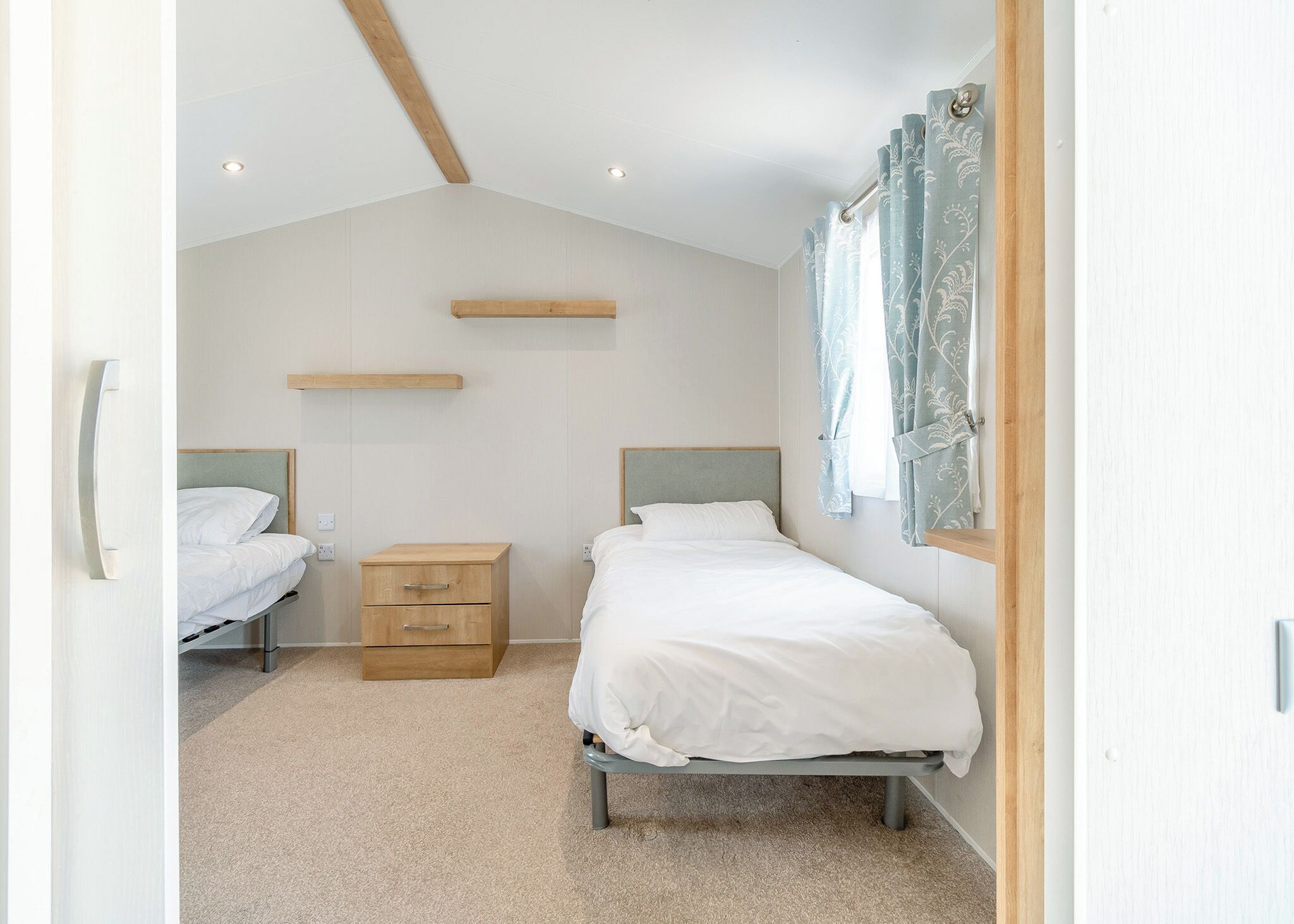 Un dormitorio con dos camas, una mesita de noche de madera, una estantería empotrada y una ventana con cortinas.