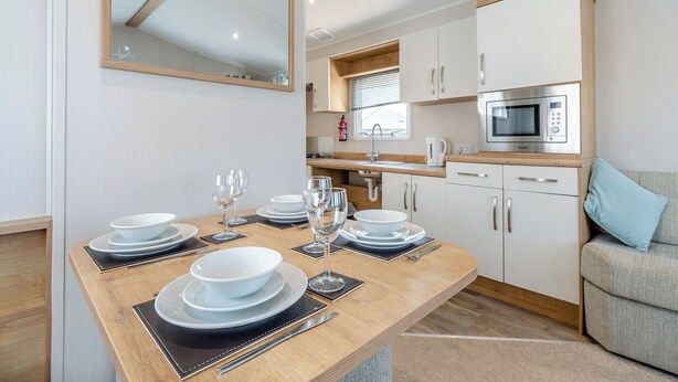 A modern kitchen with white cabinets, a microwave, and a dining table set with plates and glasses. - Gold Caravan 6 WCF (Pet) (Skegness)