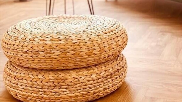 Un pouf rond en osier dans un salon doté d’un plancher en bois, une petite table ronde, un cactus et un canapé avec un coussin.