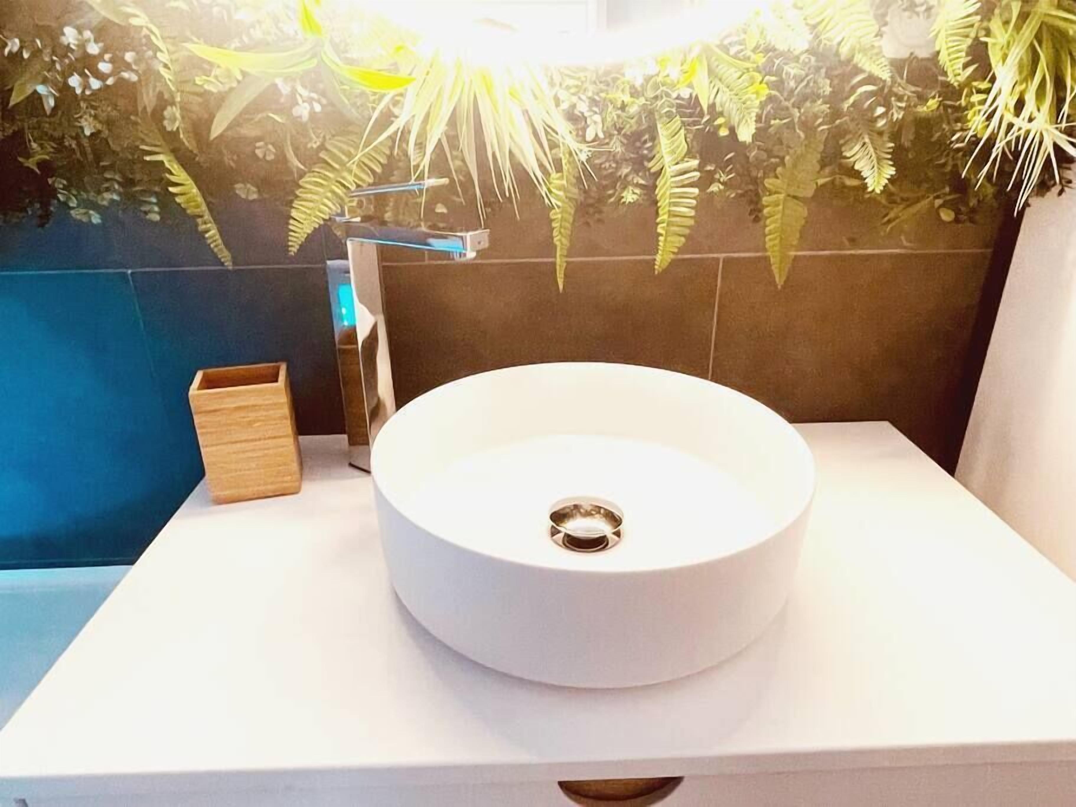 A bathroom with a round white sink, a wooden box, and a wall decorated with green plants.