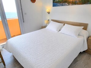 Une chambre avec un grand lit, une table de chevet, une lampe fixée au mur et une vue sur la mer, visible à travers une porte vitrée.