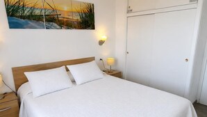Un lit bien fait, recouvert d’une couvre-lit blanche, avec deux oreillers, une tête de lit en bois, une lampe de chevet et une table de chevet dotée d’un tiroir.