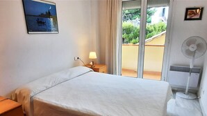 Une chambre d’hôtel comprenant un lit, une table de chevet, une lampe, une fenêtre donnant sur un espace verdoyant et un ventilateur sur pied.