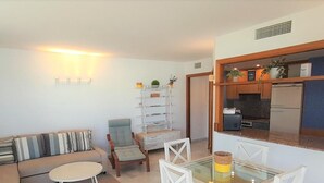 Un espace de vie compact comprenant un canapé, une table à manger avec un plateau en verre et des chaises, un petit coin cuisine avec un réfrigérateur et un meuble d’étagères agrémenté d’objets décoratifs.