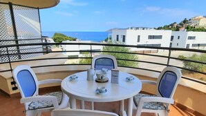 Un balcon avec une table ronde et quatre chaises, une enseigne indiquant « Sucre », et une vue sur la mer et les bâtiments en arrière-plan.