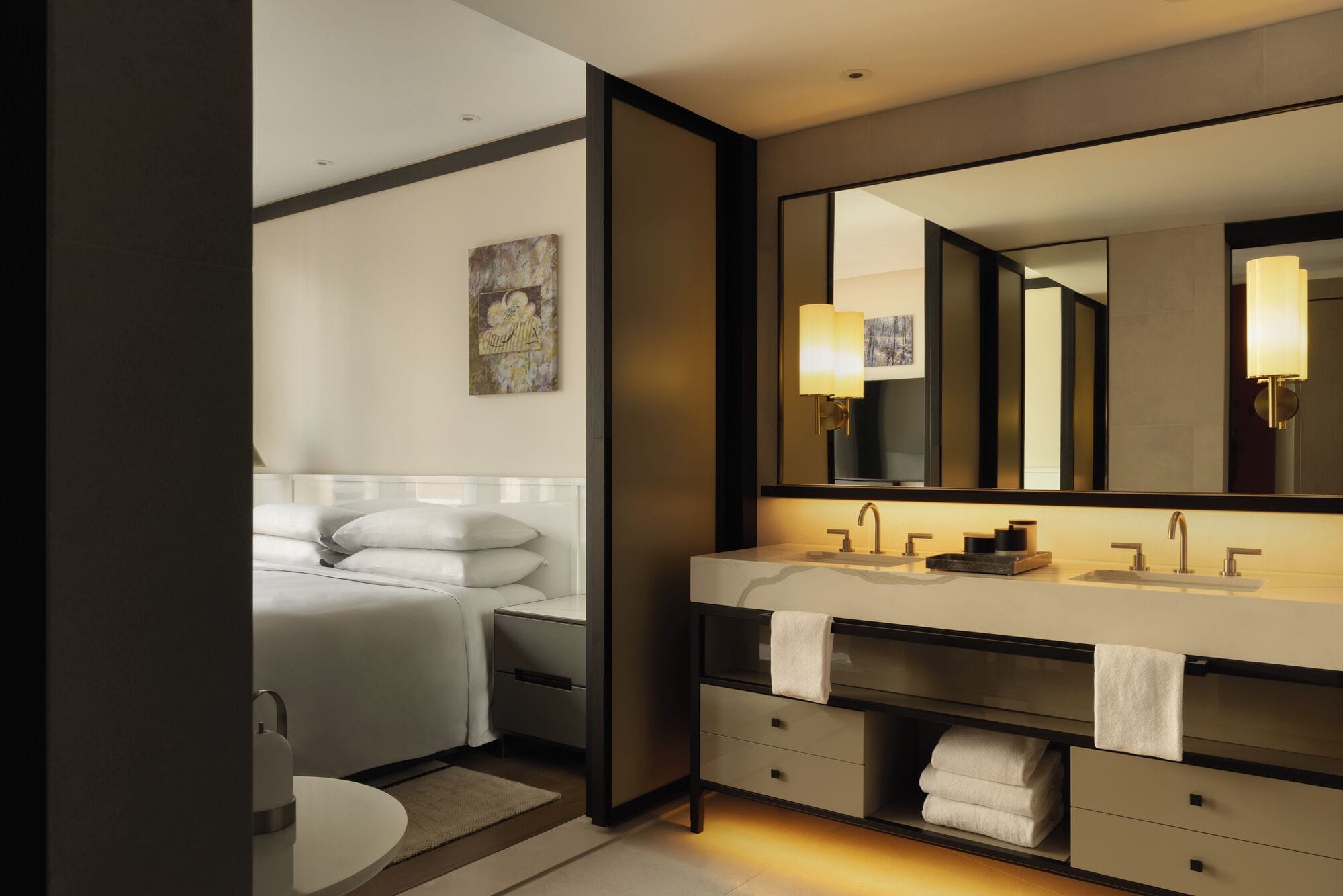 Un baño moderno con un espejo grande, tocador con lavamanos y una cama con almohadas.