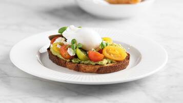 A plate with a sandwich topped with avocado, tomato, and a poached egg, with a bowl of croissants in the background.
