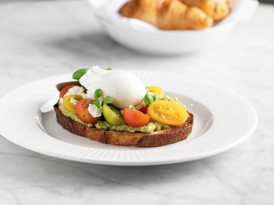 A plate with a sandwich topped with avocado, tomato, and a poached egg, with a bowl of croissants in the background.