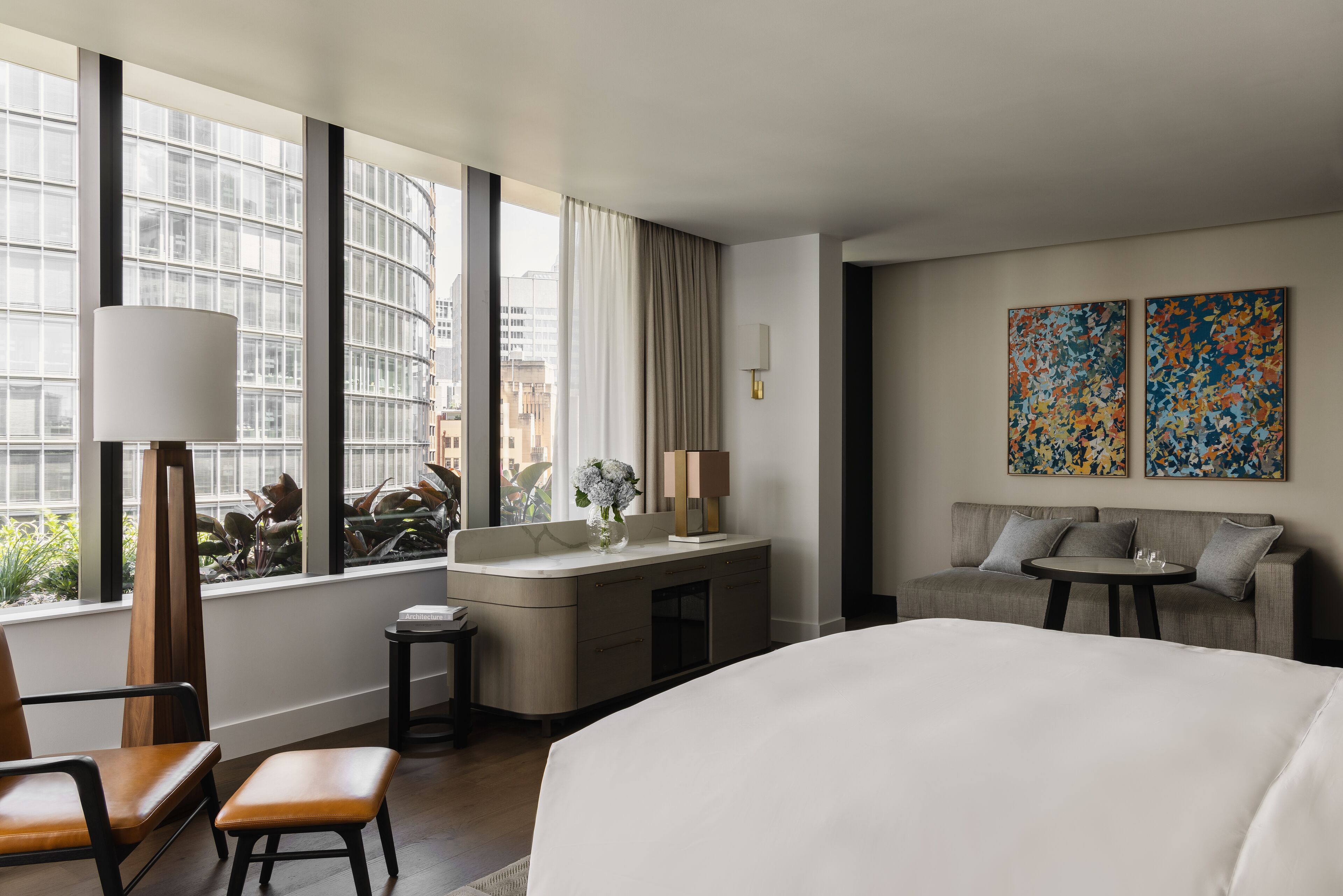 A hotel room with a large bed, a desk with a lamp, a sofa, and a view of the city through large windows.