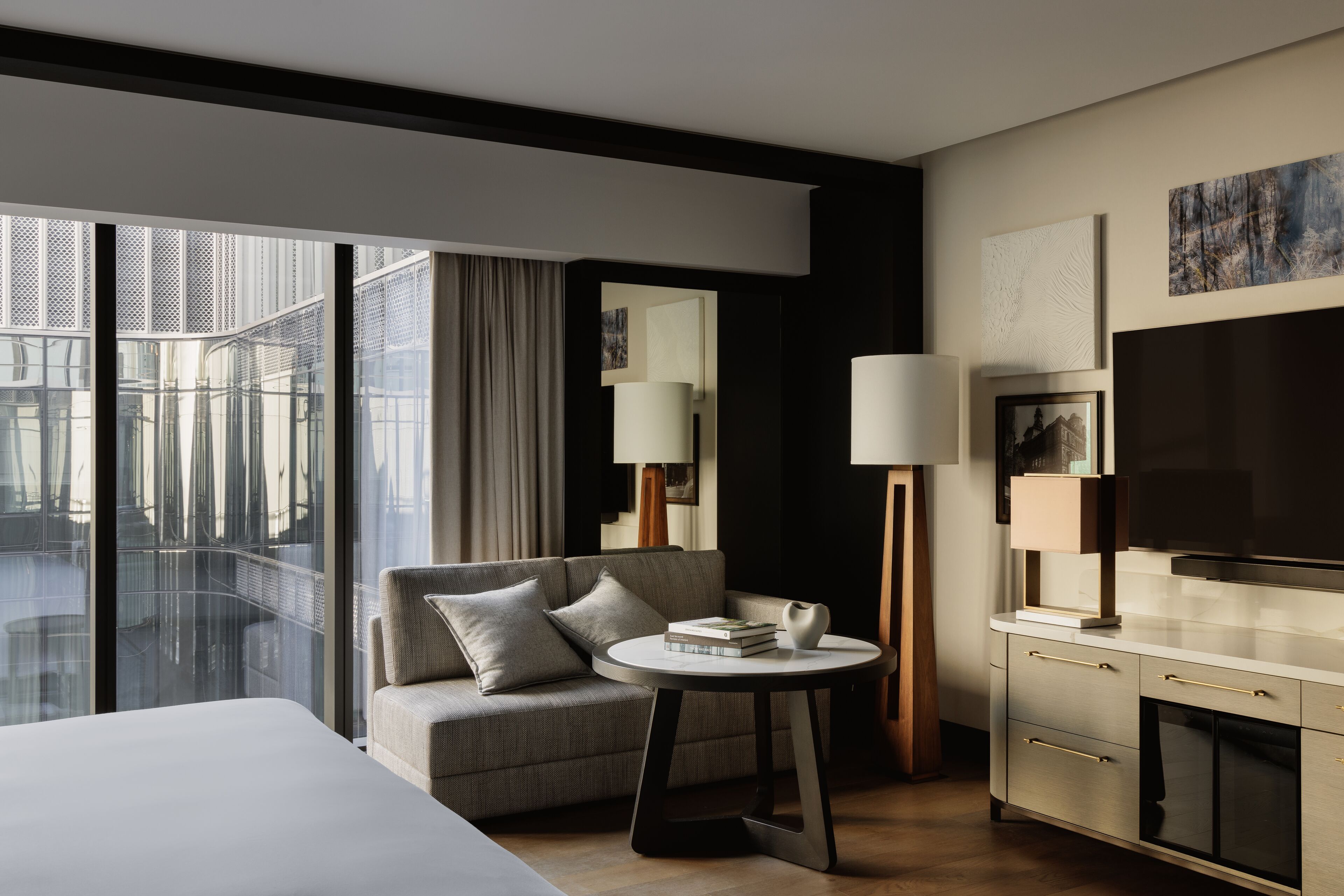 Una habitación de hotel moderna con una cama grande, un sofá, una mesita redonda con libros, un televisor y un balcón con vistas a la ciudad.