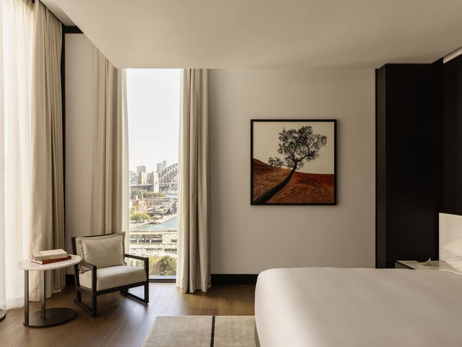 A modern hotel room with a large bed, a chair, a small table, and a city view through large windows.
