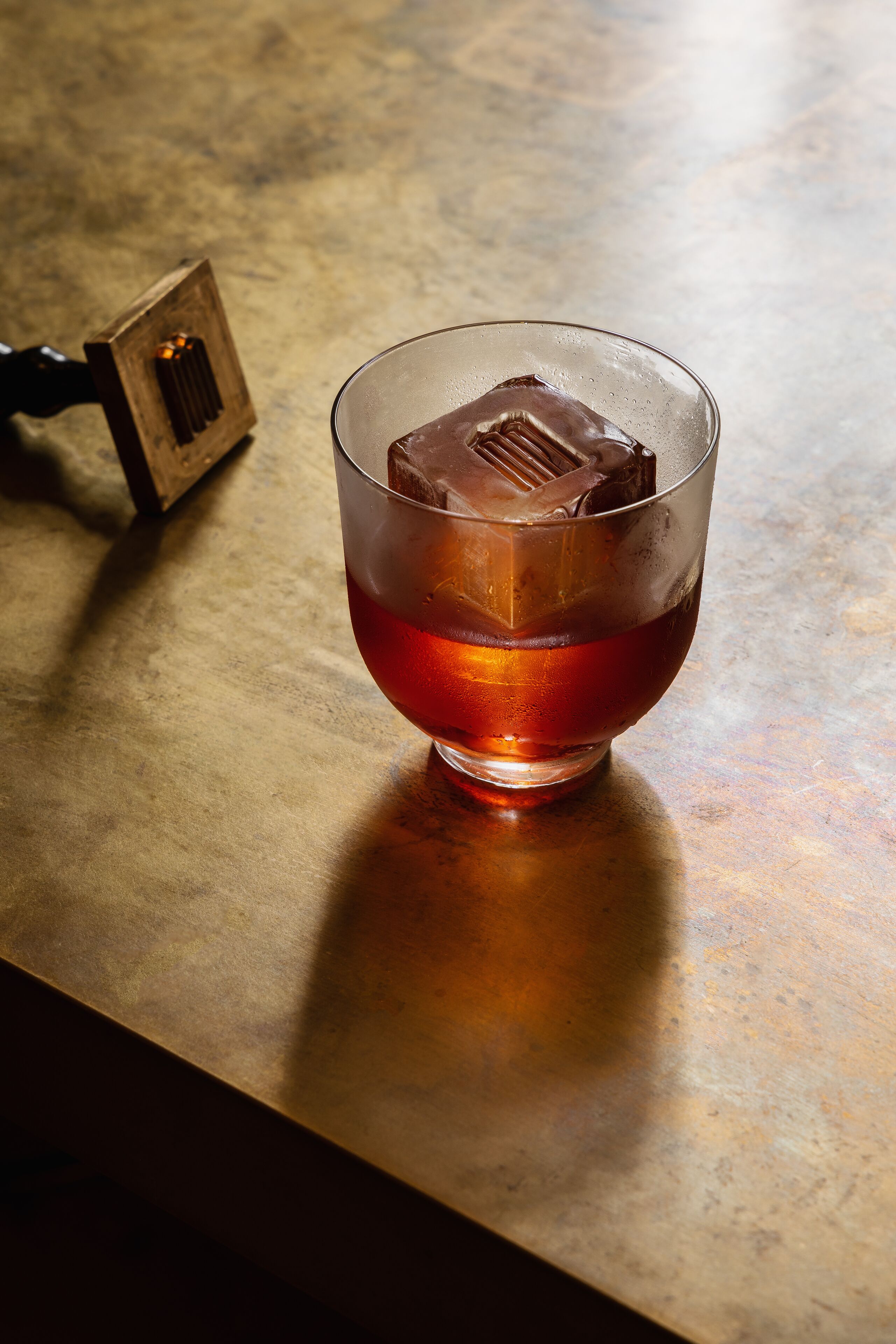 Un vaso con líquido oscuro y cubitos de hielo sobre una superficie rugosa.