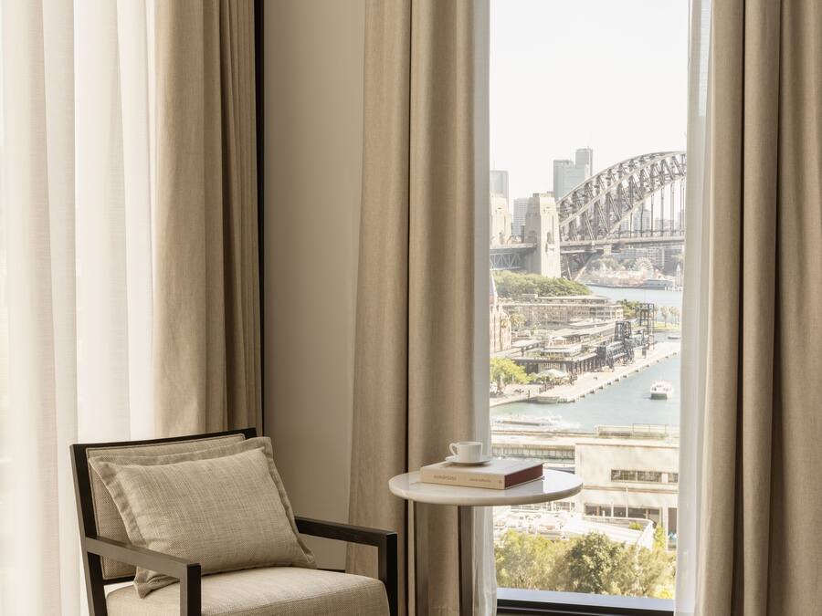 A room with a view of a bridge and cityscape, a chair, and a small table with a book.