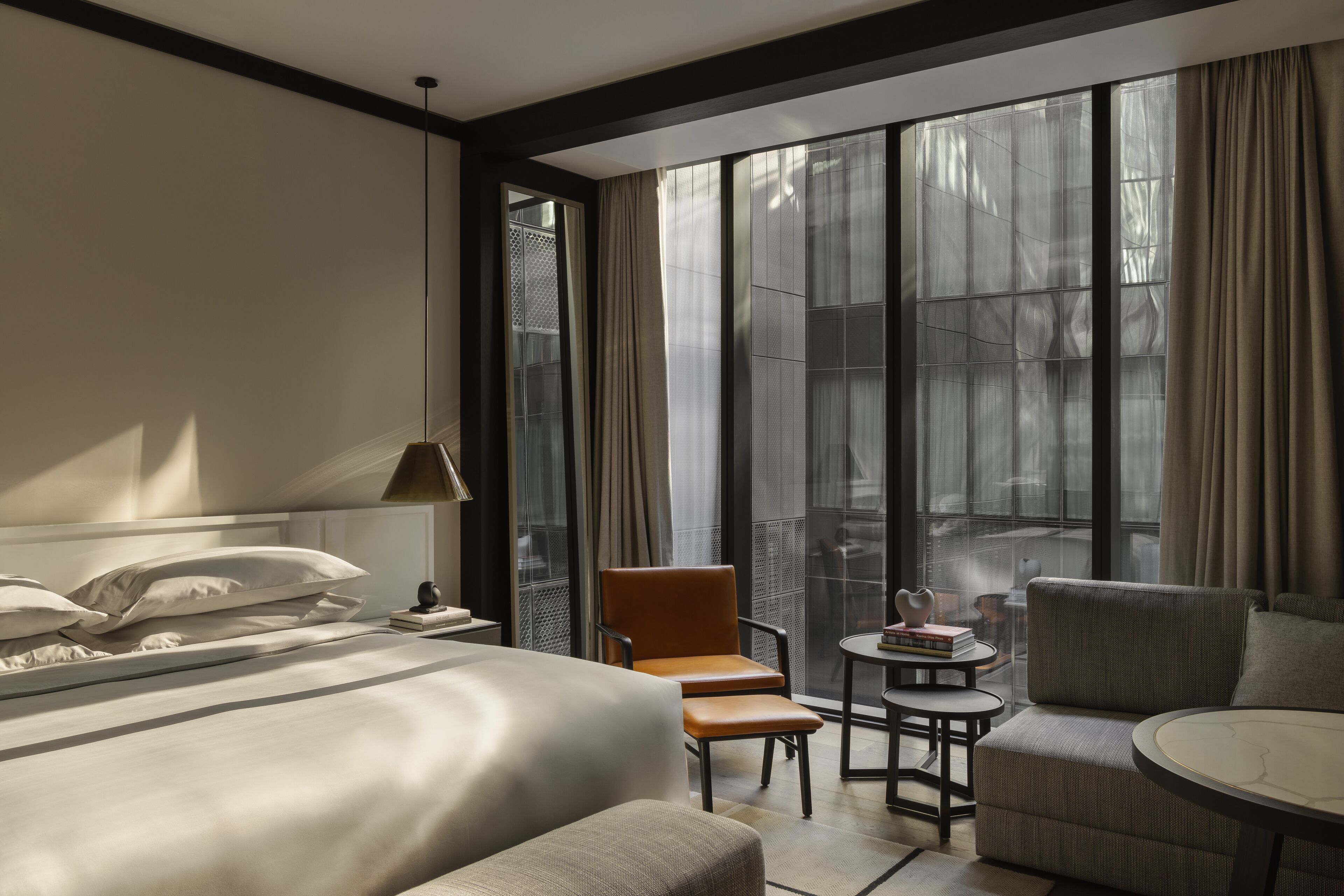 A modern hotel room with a large bed, a sitting area with a chair and a small table, and a large window with curtains.